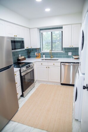 Una cocina moderna con armarios blancos, electrodomésticos de acero inoxidable, un ventanal con persianas y una alfombra color beige.