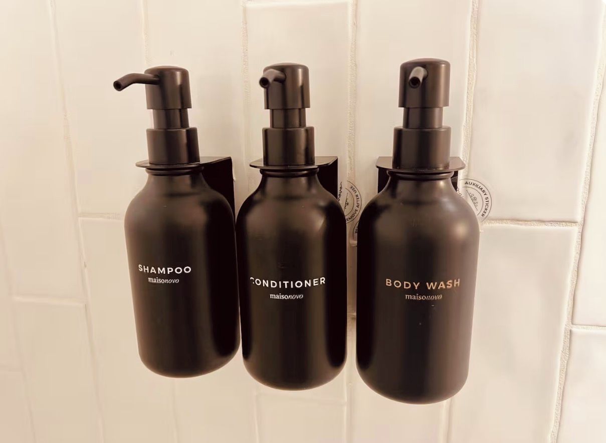 Three black bottles with pump dispensers labeled 'SHAMPOO,' 'CONDITIONER,' and 'BODY WASH' against a white tiled wall.