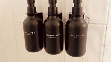 Three black bottles with pump dispensers labeled 'SHAMPOO,' 'CONDITIONER,' and 'BODY WASH' against a white tiled wall.