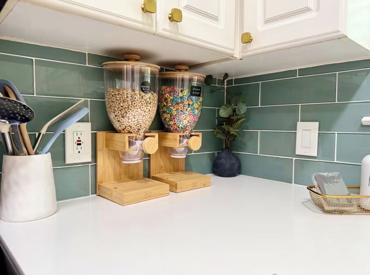 A kitchen counter with two glass jars containing cereal, a white countertop, and a green tiled backsplash.