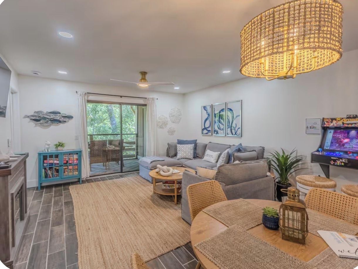 A modern living room with a sofa, coffee table, and a dining area. There is a ceiling fan, a large pendant light, and a television.