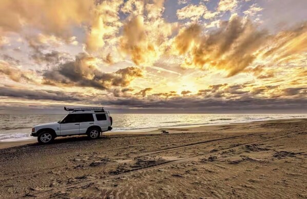Ein weißer SUV parkt auf einem Sandstrand, im Hintergrund ein dramatischer Sonnenuntergang.