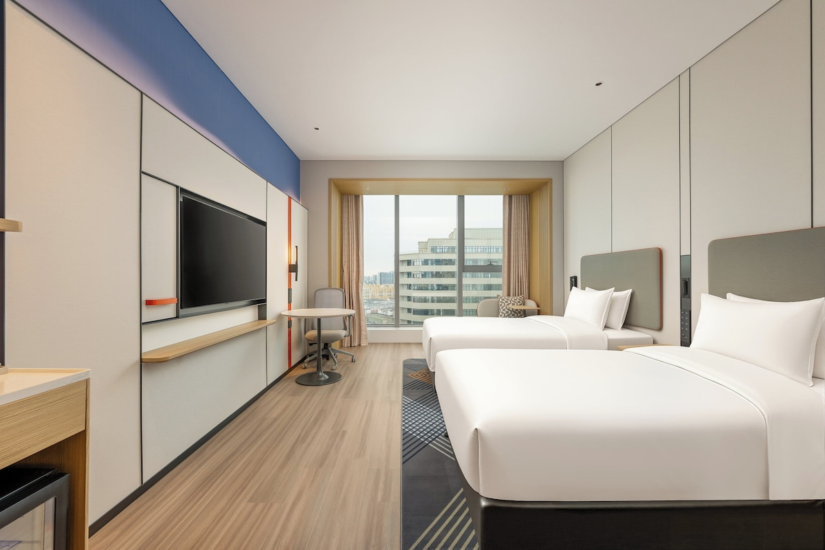 Habitación de hotel con dos camas, televisión, escritorio y ventana grande con vista a la ciudad.