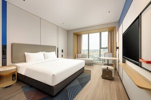 A modern hotel room with a large bed, a desk, a chair, a TV, and a view of the city.