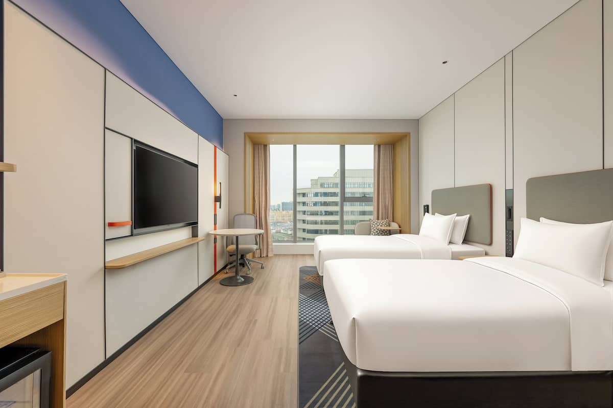 Habitación de hotel con dos camas, televisión, escritorio y ventana grande con vista a la ciudad.