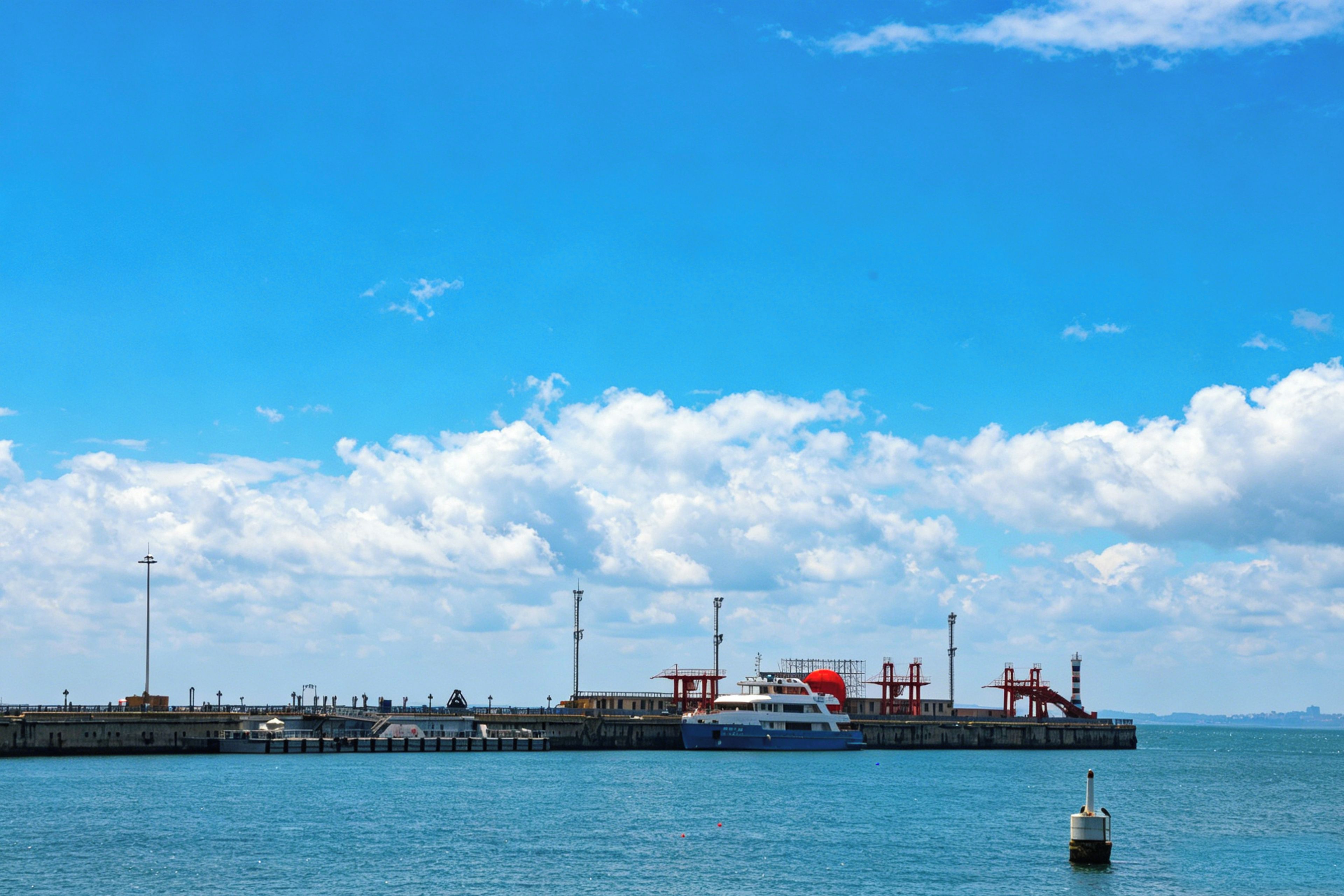 A large industrial port with a ship docked, a pier with structures, and a clear blue sky.