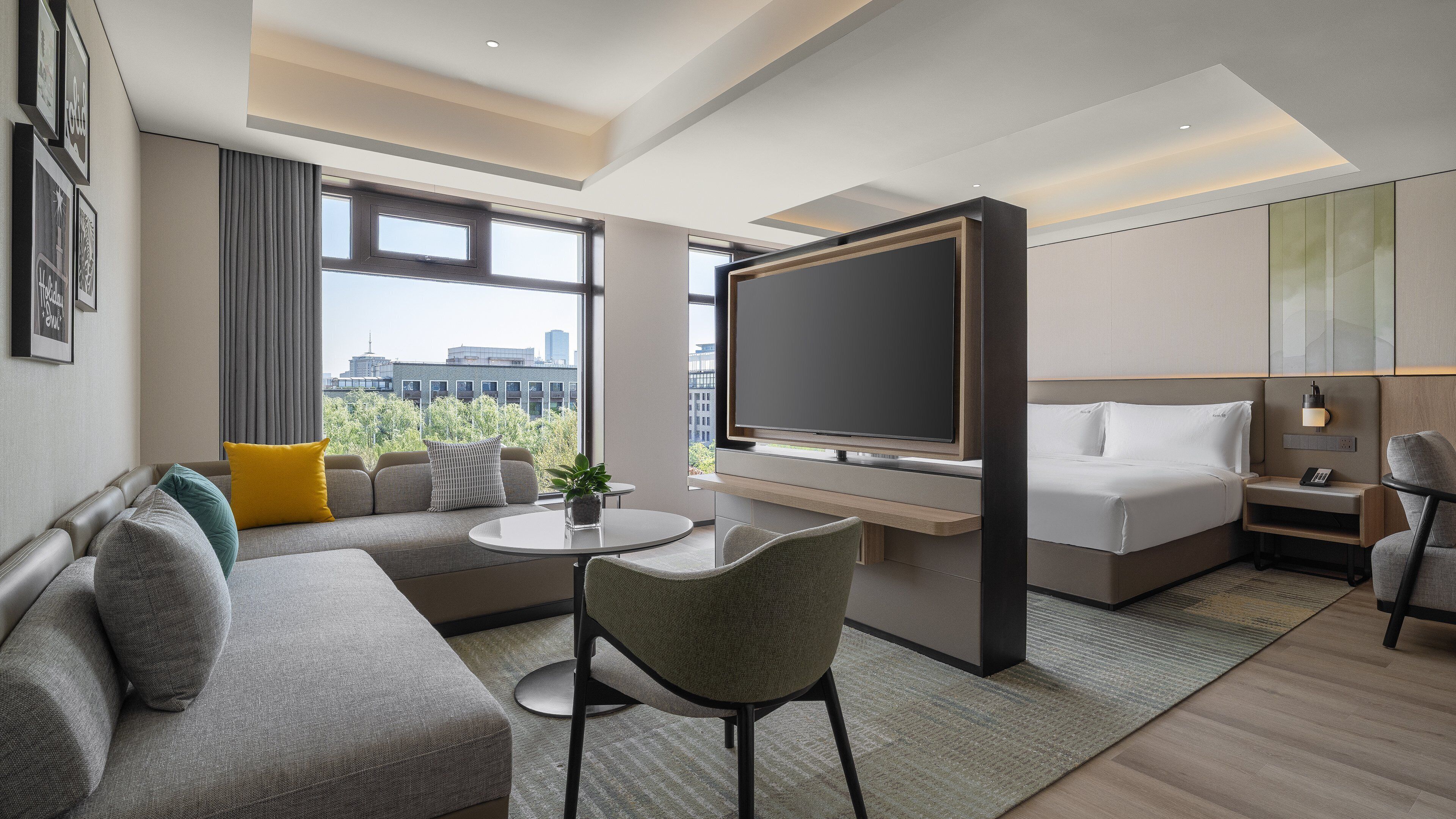 A modern hotel room with a sofa, a round table, a TV, a bed, and a city view.