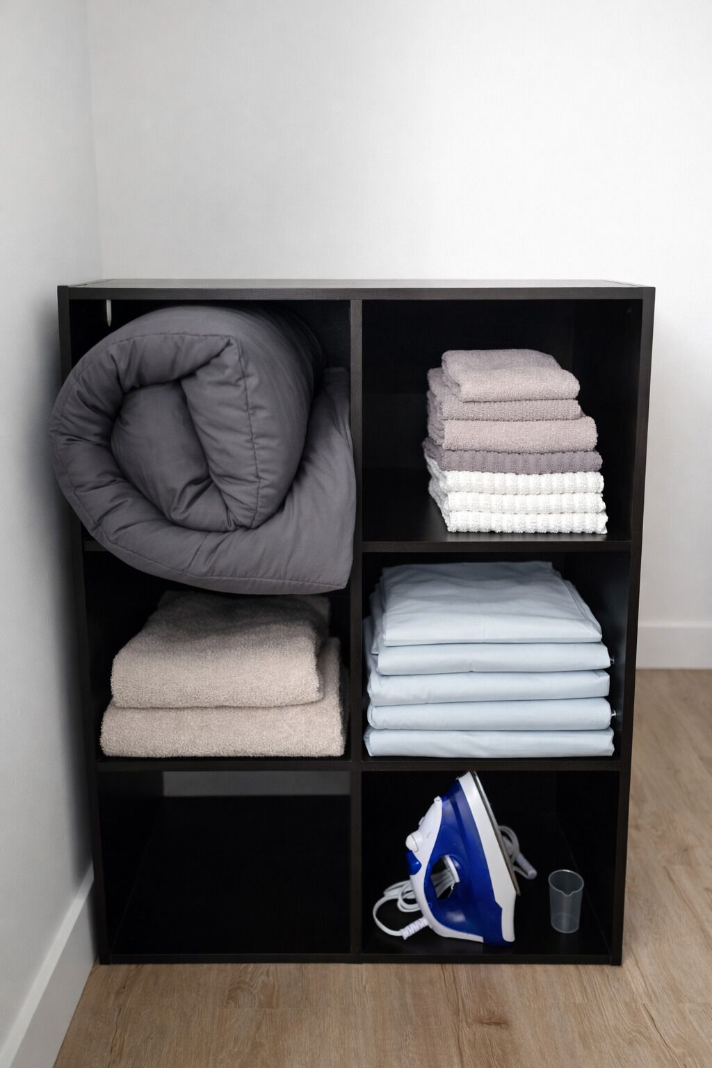 A black storage unit with a grey cushion, folded towels in various shades, and a blue iron.
