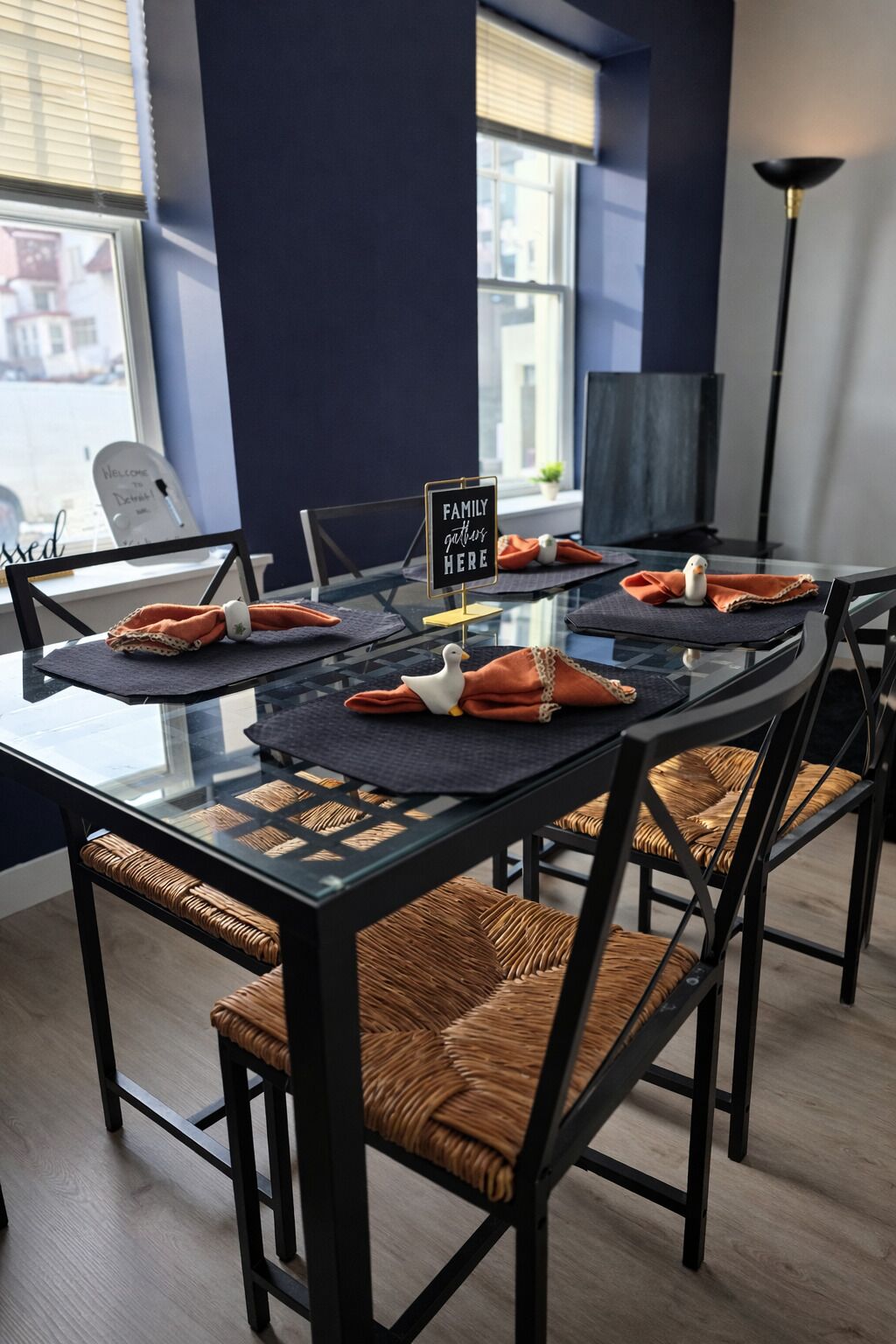 A modern dining area with a glass table, black metal chairs, and orange napkins. A sign reads "Family Gather Here."