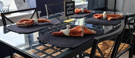 A modern dining area with a glass table, black metal chairs, and orange napkins. A sign reads "Family Gather Here."
