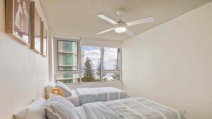 Ein Schlafzimmer mit zwei Betten, einem Deckenventilator, einem Fenster mit Blick auf Bäume und gerahmten Bildern an der Wand.