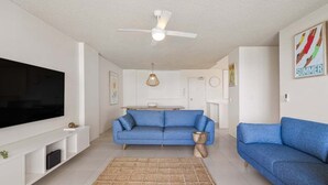 Ein modernes Wohnzimmer mit Flachbildfernseher, zwei blauen Sofas, einem Couchtisch, einem Deckenventilator und einem gewebten Teppich.
