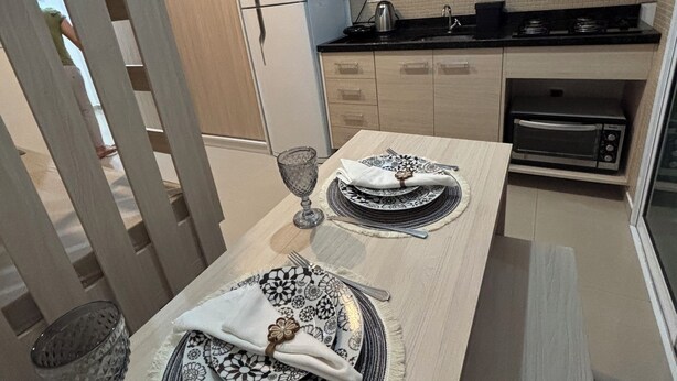 A modern kitchen with light wood cabinets, a microwave, and an oven. There is a dining table set with plates, cutlery, and a glass.