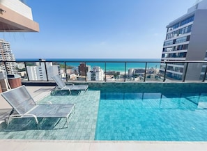 A rooftop pool with lounge chairs, a view of the ocean, and surrounding buildings.
