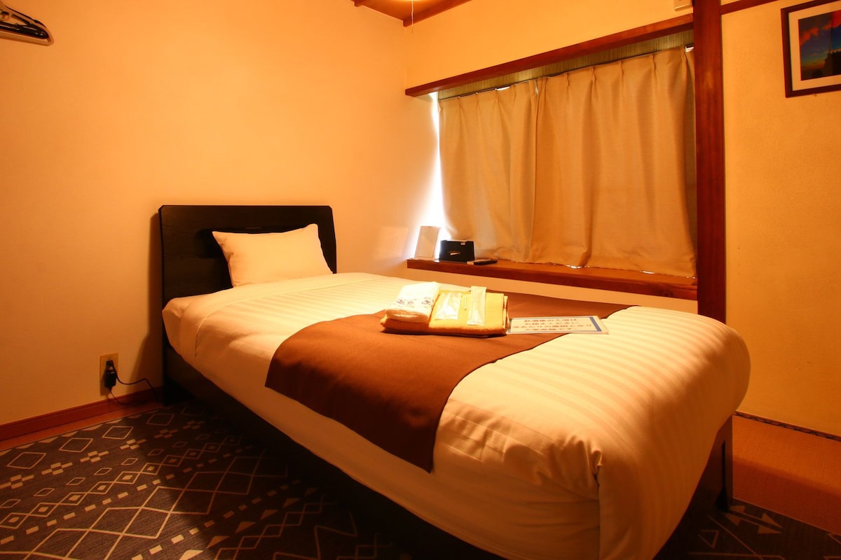 Habitación de hotel con una cama individual, una ventana con cortinas, una mesita de noche con lámpara y un suelo estampado.