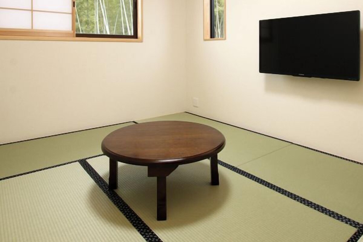 Ein Zimmer mit Tatami-Böden, einem runden Tisch, einem an der Wand befestigten Fernseher und einem Fenster mit Blick auf Bäume.