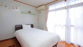 A bedroom with a bed, wooden floor, white walls, a window with sheer curtains, and a wall-mounted air conditioner.