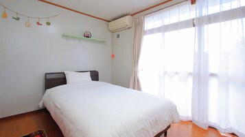 Quarto com cama, piso de madeira, paredes brancas, janela com cortinas leves e ar-condicionado fixado na parede.