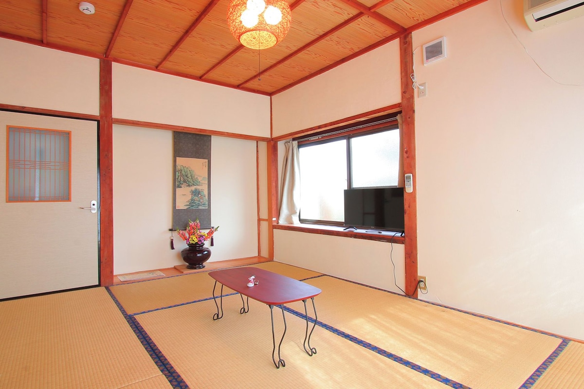 Una habitación japonesa tradicional con suelo de tatami, puertas correderas, una pequeña mesa, un jarrón con flores y una ventana con vistas.