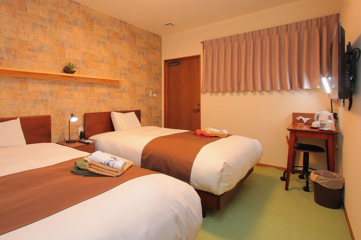 Habitación de hotel con dos camas, cabecero de madera con estante, escritorio con silla, televisión y ventana con cortinas.