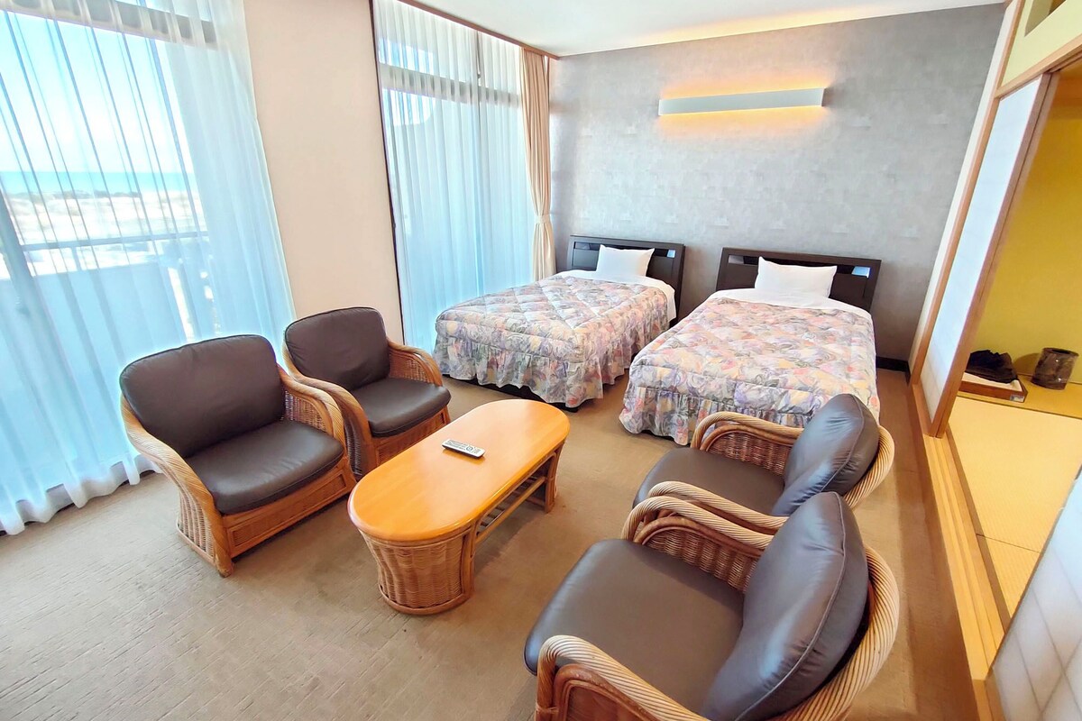Ein Hotelzimmer mit zwei Betten, einem Schlafsofa, zwei Sesseln, einem kleinen Tisch und Meerblick durch große Fenster.