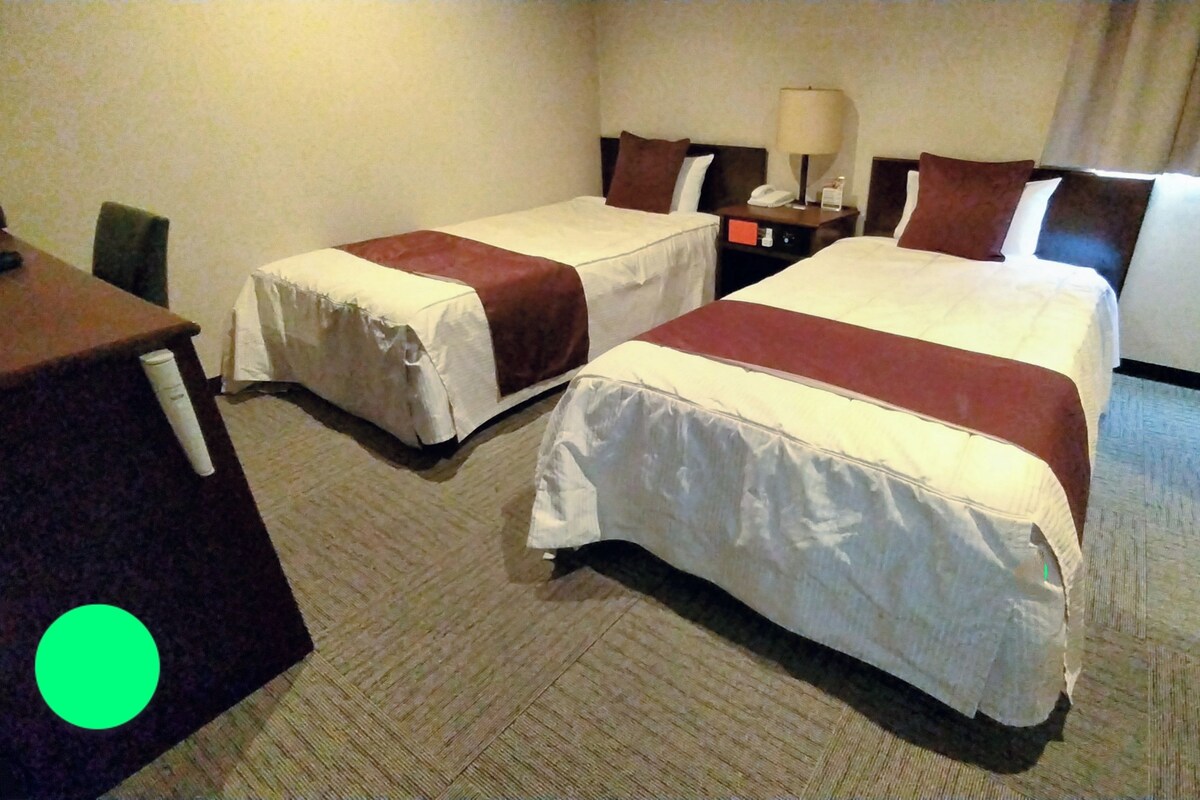 A hotel room with two beds, a desk, a chair, and a nightstand with a lamp.