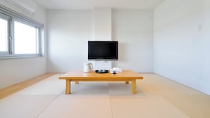 Een minimalistisch ingerichte kamer met tatami vloeren, een houten tafel met waterkoker en theeservies, en een flatscreen televisie aan de muur.