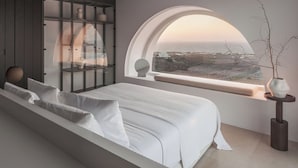 Ein modernes Schlafzimmer mit einem großen Bett, einer Glastausch und Blick auf die Küste durch ein großes Rundbogenfenster.