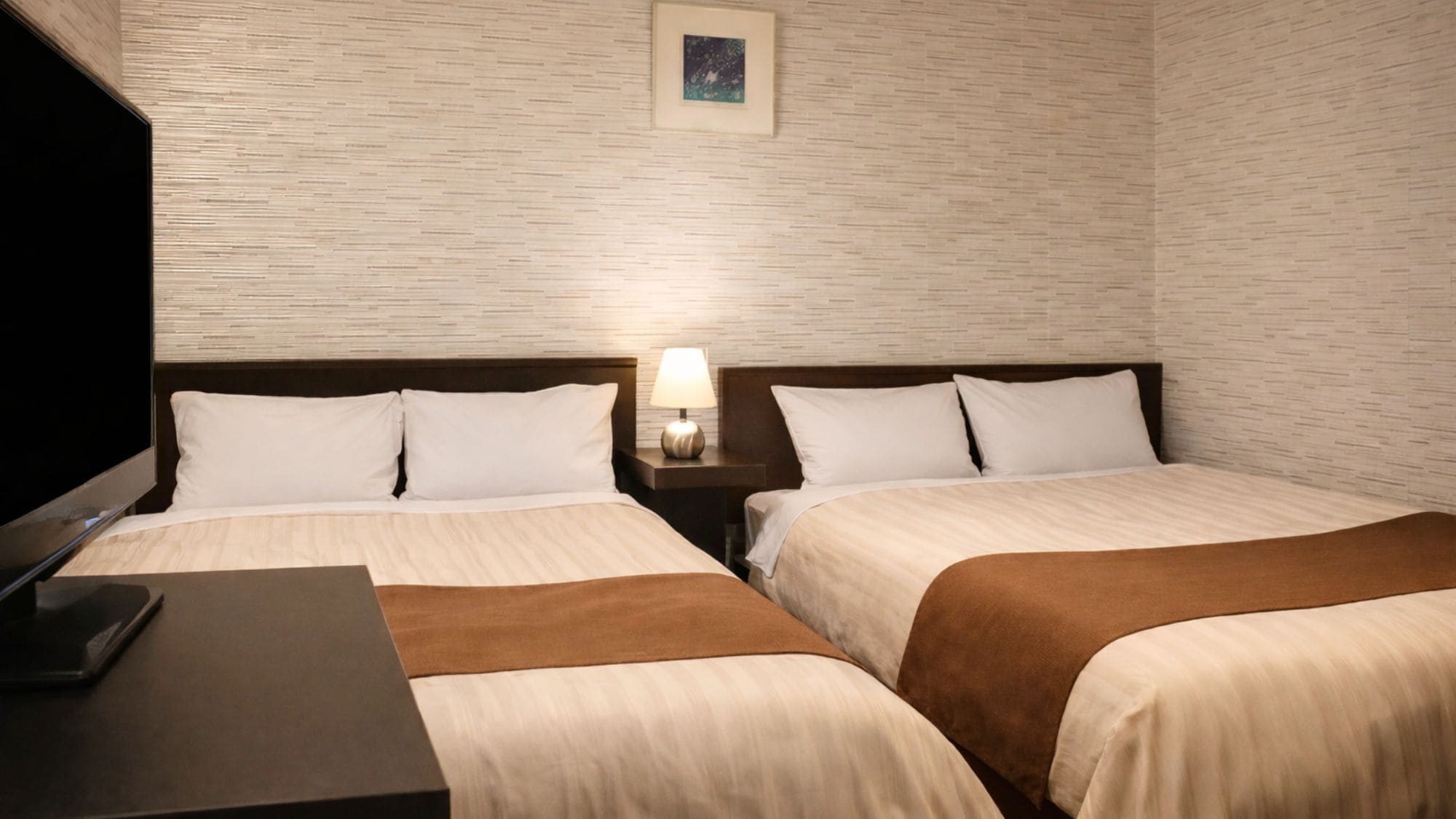 Ein Hotelzimmer mit zwei Betten, einem Fernseher, einem Nachttisch mit Lampe und einem gerahmten Bild an der Wand.