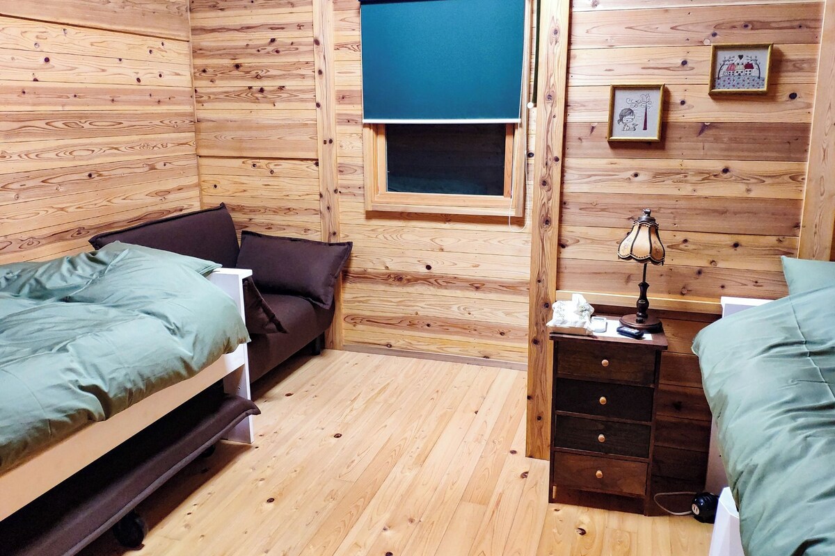 A cozy cabin-style room with wooden walls, a window with a blue blind, a small desk with a lamp, and two beds with green bedding.