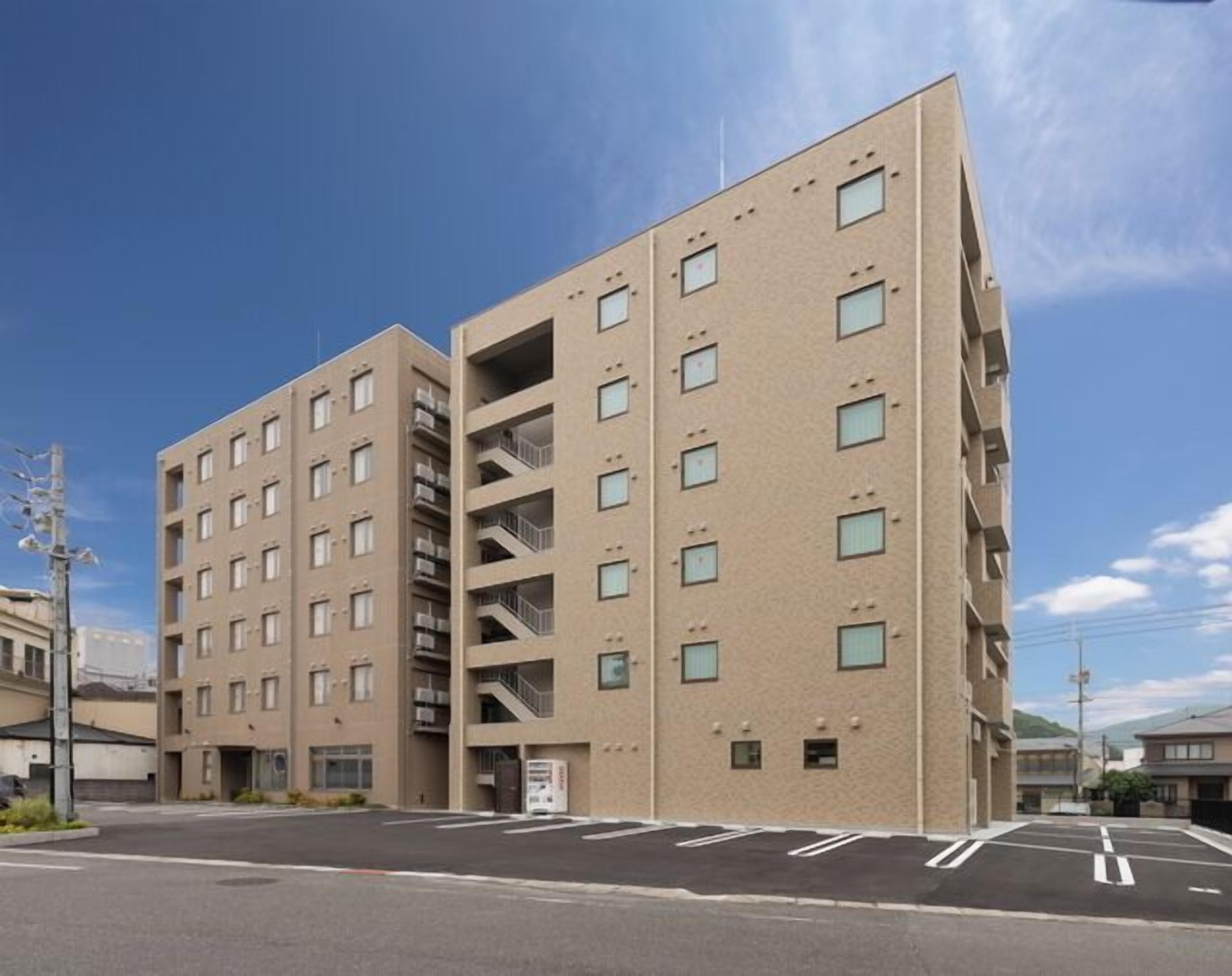 A modern multi-story building with a beige facade, multiple balconies, and a parking lot in front.