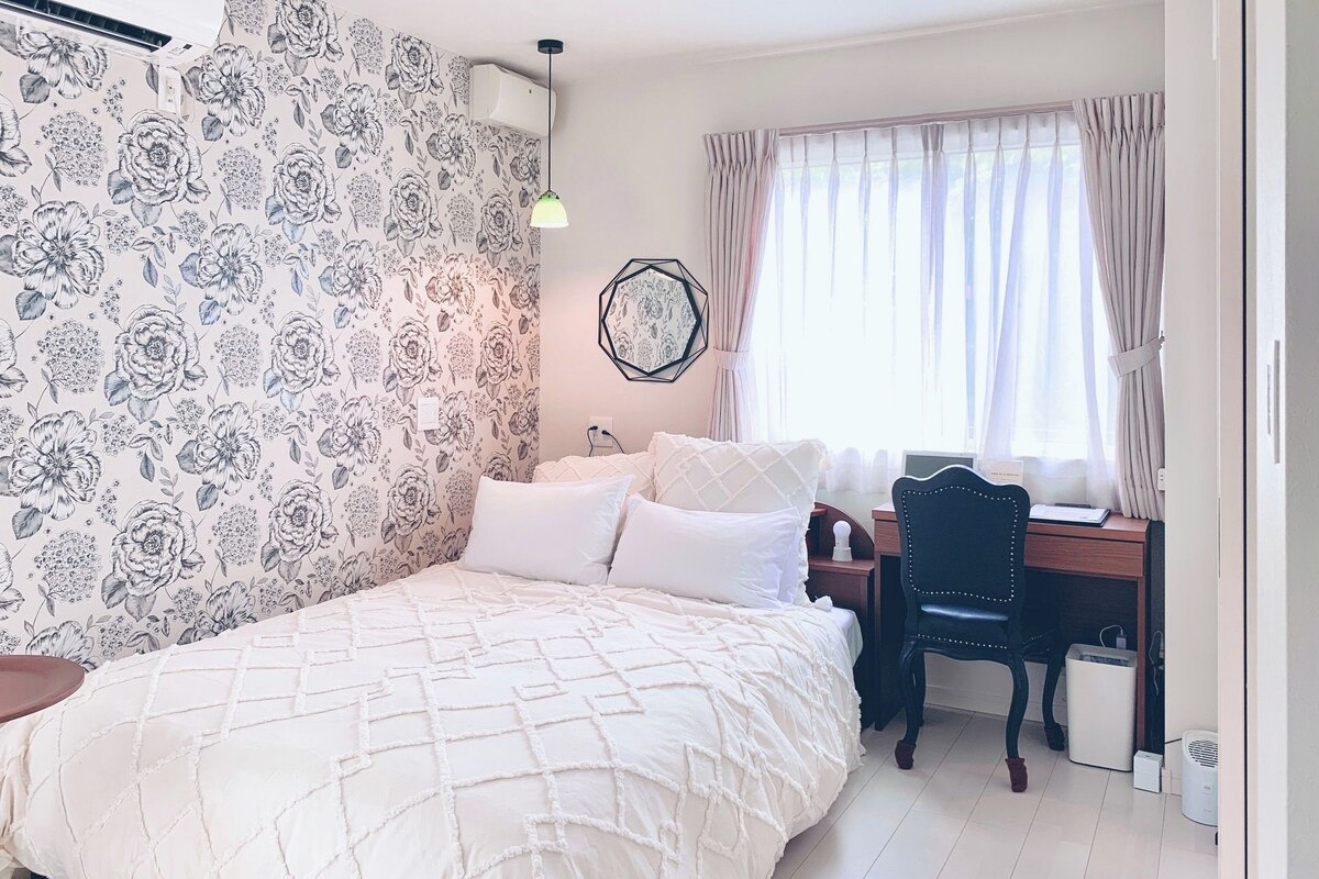 A bedroom with a bed, a desk, and a chair. There is a window with curtains, a wall-mounted light, and a wall decoration.