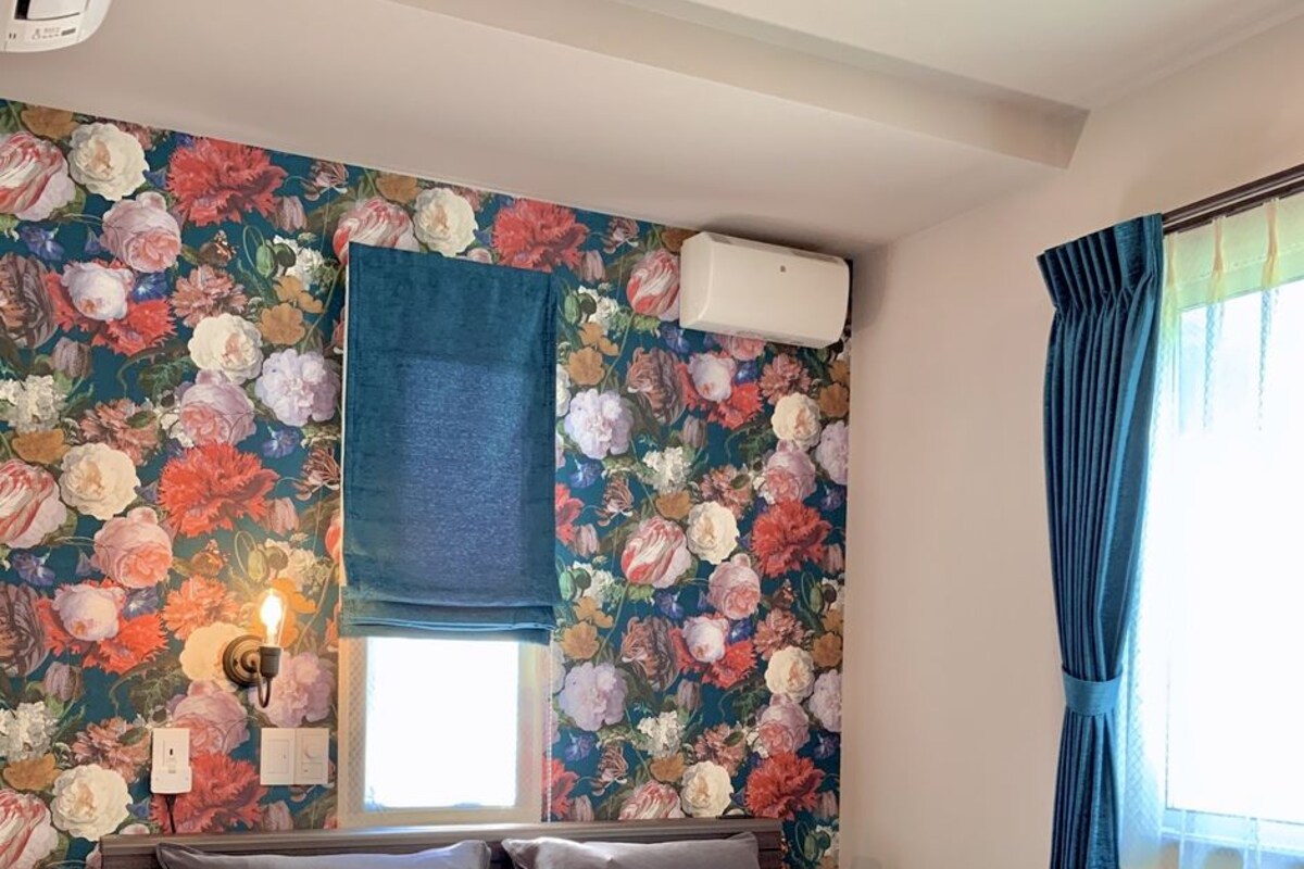 A bedroom with a floral wallpaper, a bed with grey bedding, a nightstand, a wall-mounted lamp, and a window with blue curtains.