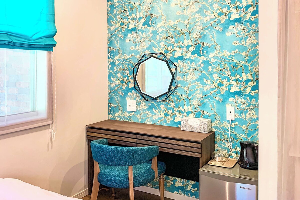 A bedroom with a bed, a desk, a chair, a mirror, and a coffee machine.