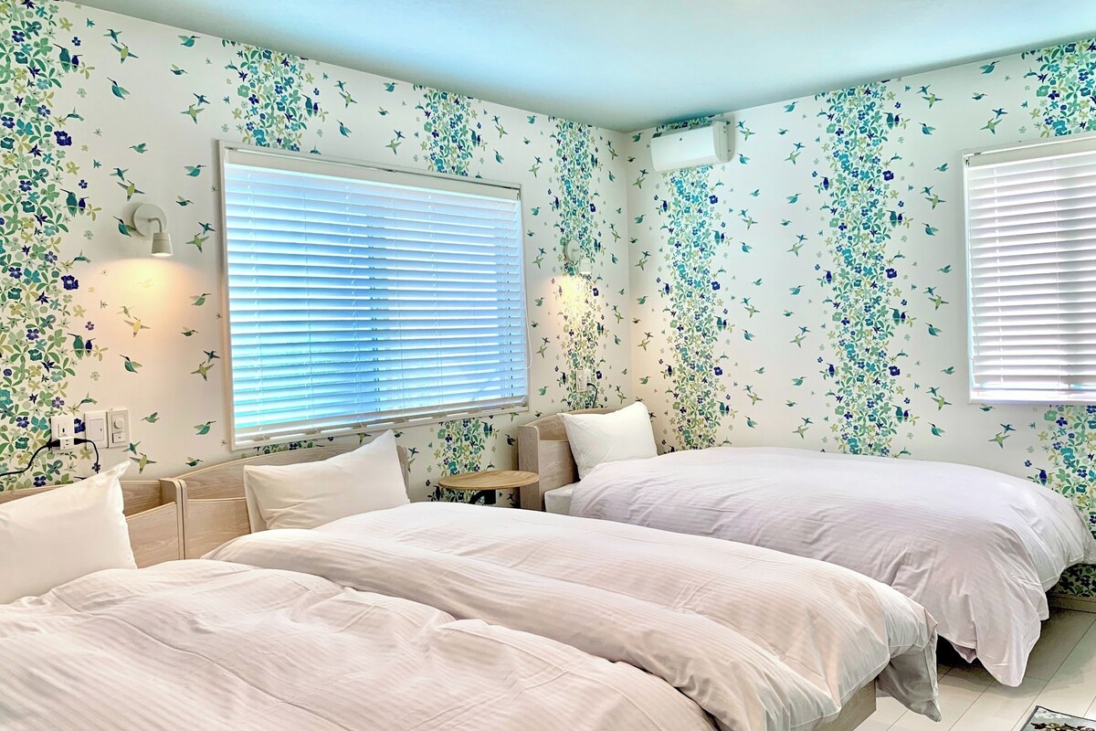 A bedroom with two beds, a nightstand, a small table, and a window with blinds.