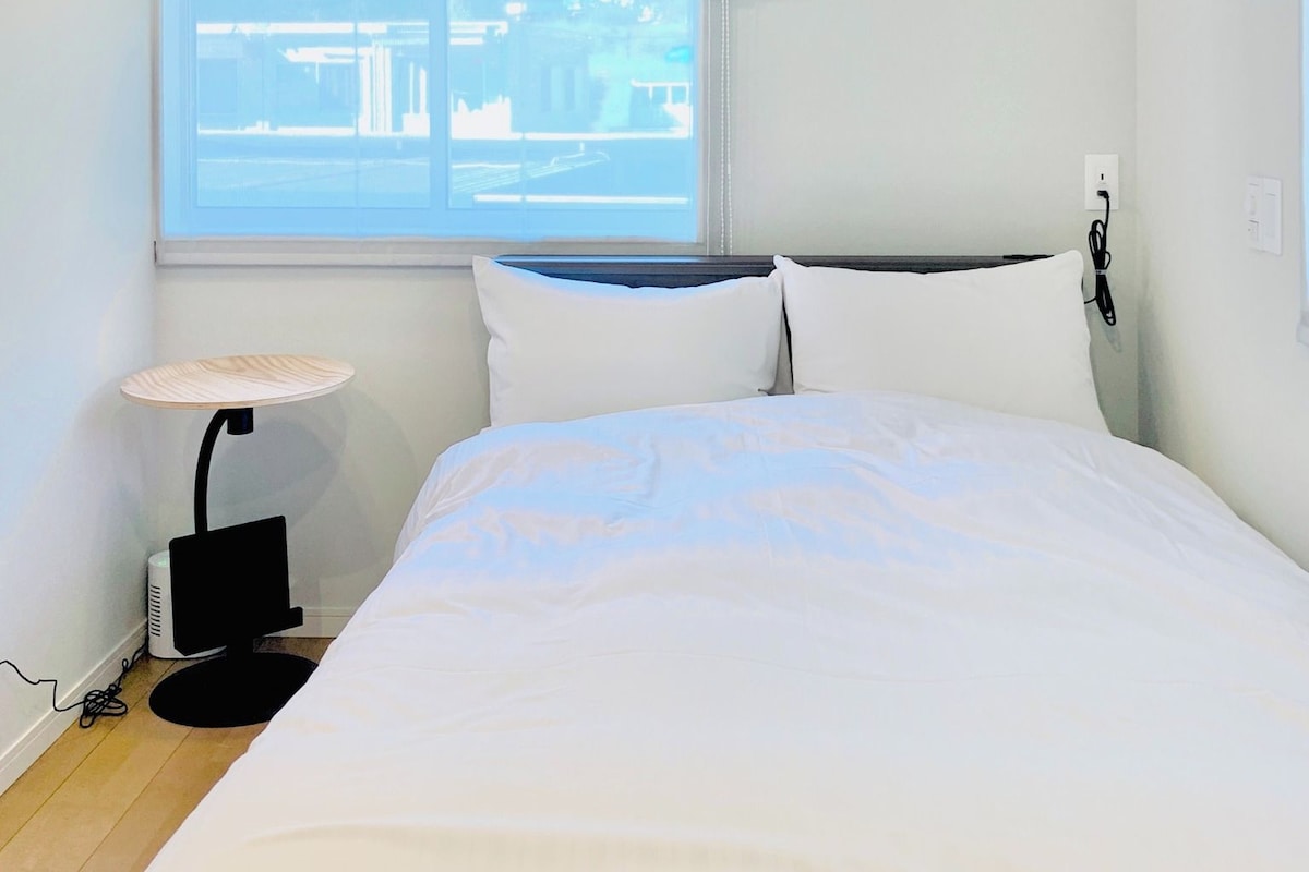 A bedroom with a bed, a bedside lamp, an air conditioner, and a window with a view of buildings.