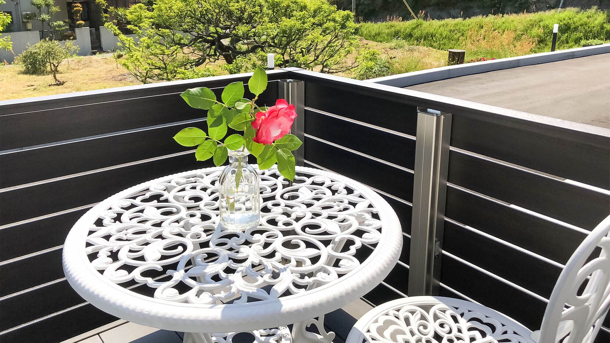 A white metal table with a glass vase and a pink rose on a balcony with a black railing.