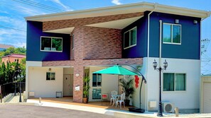 A two-story building with a brick and blue facade, a covered patio with outdoor seating, and a visible garage door. - Sparky's House / House II (Naoshima)