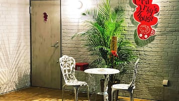 Ein Zimmer mit weißer Ziegelwand, einer Holztür, zwei weißen Stühlen, einem kleinen runden Tisch und einem Blumentopf.