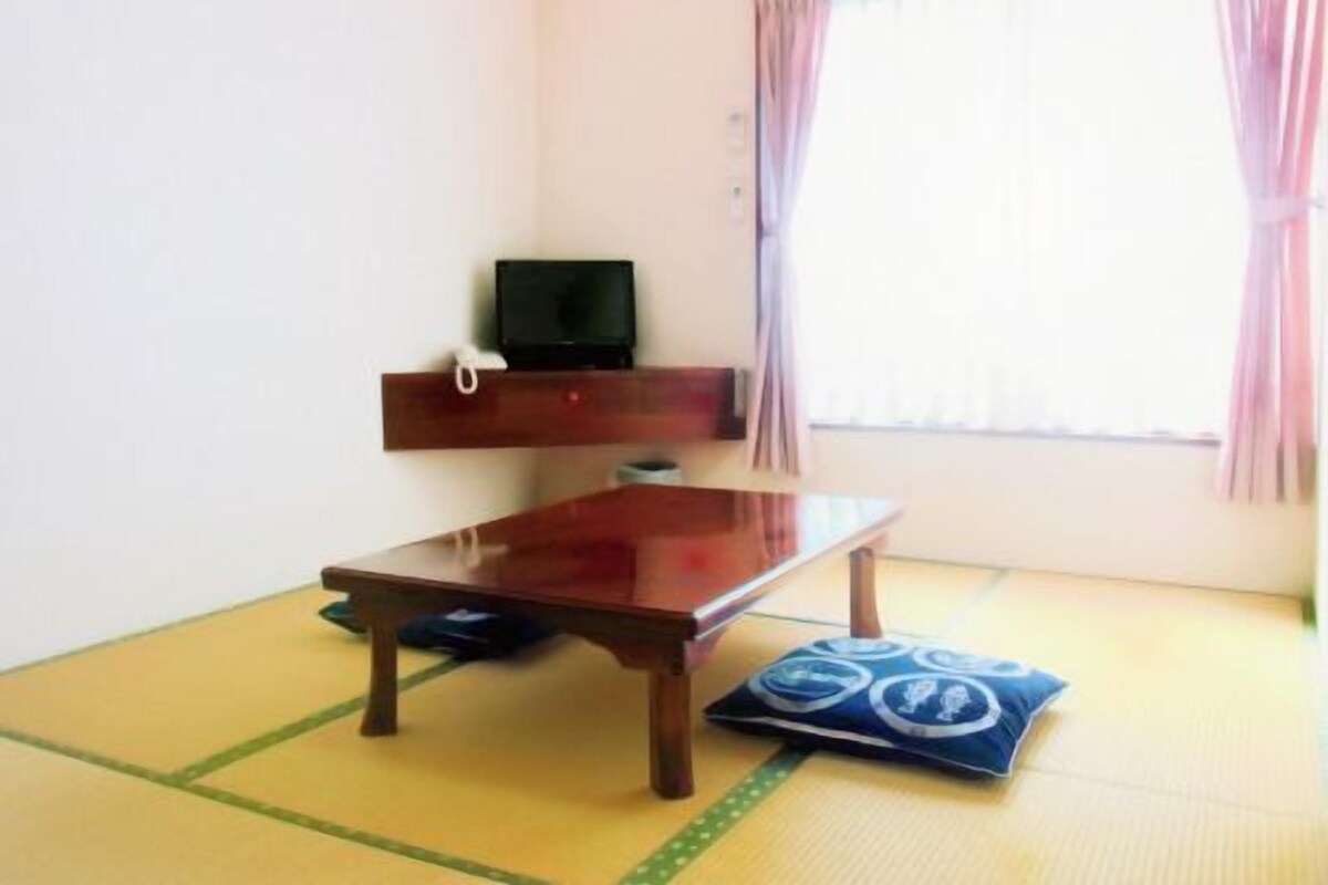 Una habitación con suelo de tatami, una mesa de madera, un televisor sobre una estantería y una ventana con cortinas.