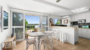 A modern kitchen with white cabinets, a dining area with white chairs, and a view of the sea through large windows. - sunny, north-facing ground-floor apartment (Noosaville)