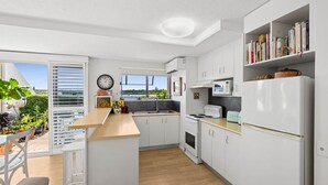 A modern kitchen with white cabinetry, a central island, a microwave, a stove, and a refrigerator. There is a window providing a view of the outdoors. - sunny, north-facing ground-floor apartment (Noosaville)