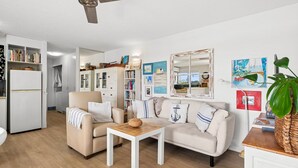 A cozy living room with a sofa, armchair, and coffee table. There is a bookshelf, a kitchen area, and artwork on the walls. - sunny, north-facing ground-floor apartment (Noosaville)