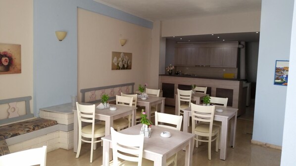 A dining area with white tables and chairs, a kitchen in the background, and a painting on the wall. - Pallada Apartment (Agios Vasileios)