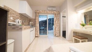 A modern kitchen with a view to a balcony, a dining area, and a bedroom with a large bed and a desk. - Pallada Apartment (Agios Vasileios)