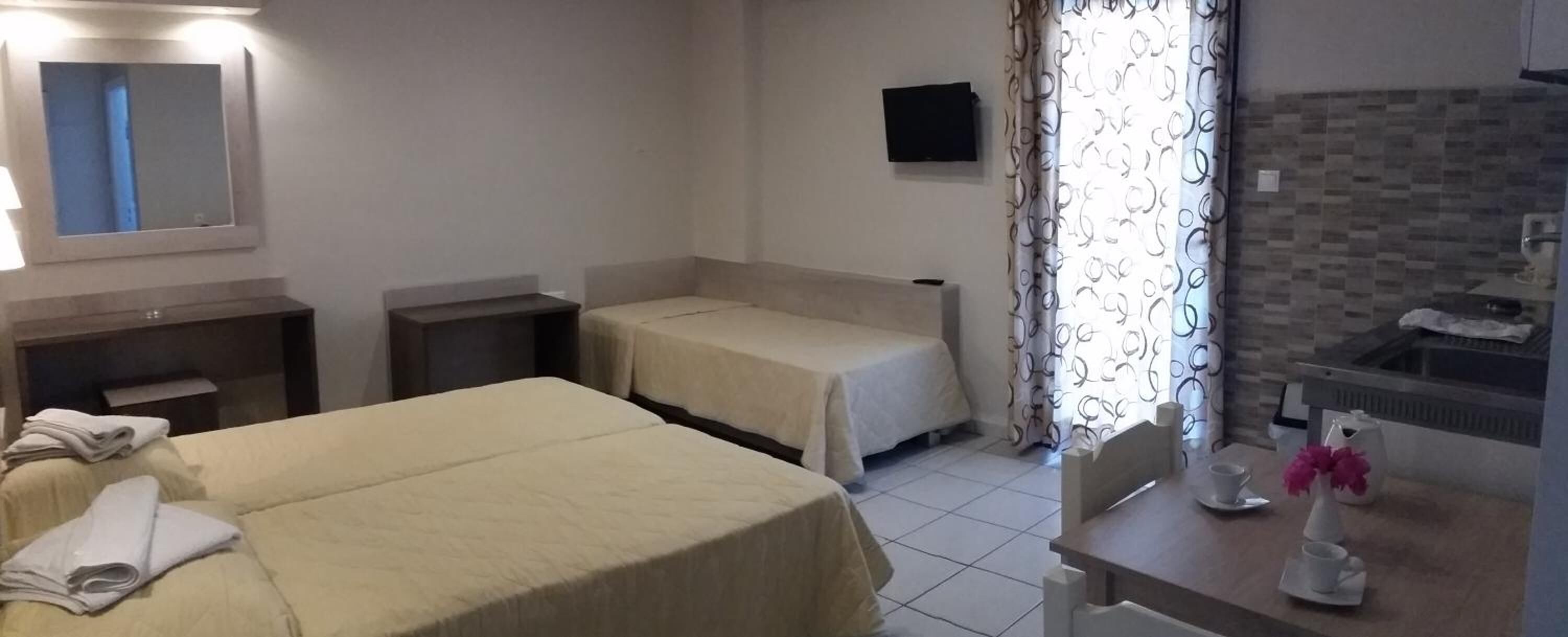 Habitación de hotel con dos camas, una mesita con un jarrón de flores, un televisor y un baño con espejo y toallero.