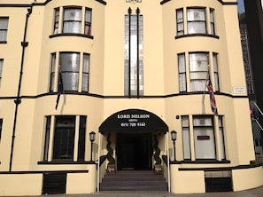Un bâtiment à la façade incurvée, doté de nombreuses fenêtres et d’une entrée imposante sur laquelle est apposée une enseigne indiquant « Hôtel Lord Nelson ».