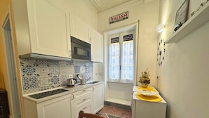 Una cocina compacta con armarios blancos, microondas y salpicadero de azulejos. Hay una pequeña mesa de comedor con una silla y un refrigerador a la derecha.