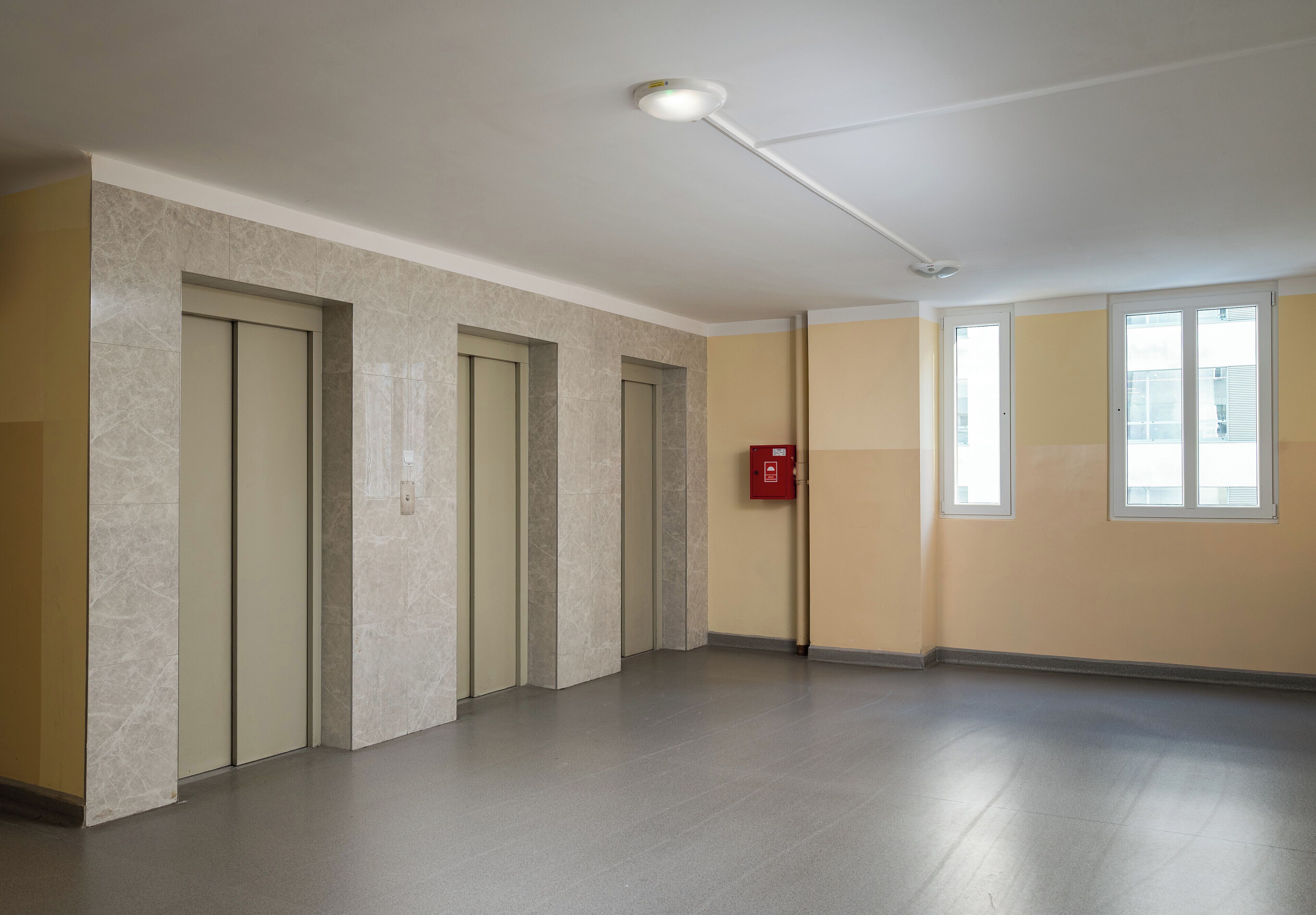 A spacious hallway with marble walls and a grey floor, leading to a door with a fire alarm.