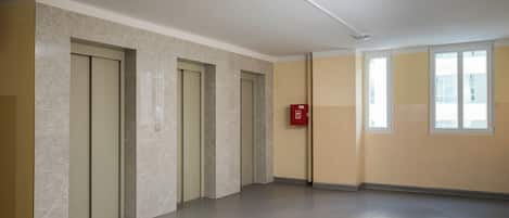 A spacious hallway with marble walls and a grey floor, leading to a door with a fire alarm.